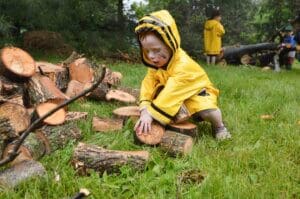 Young child in a raincoat picking up a wooden log.