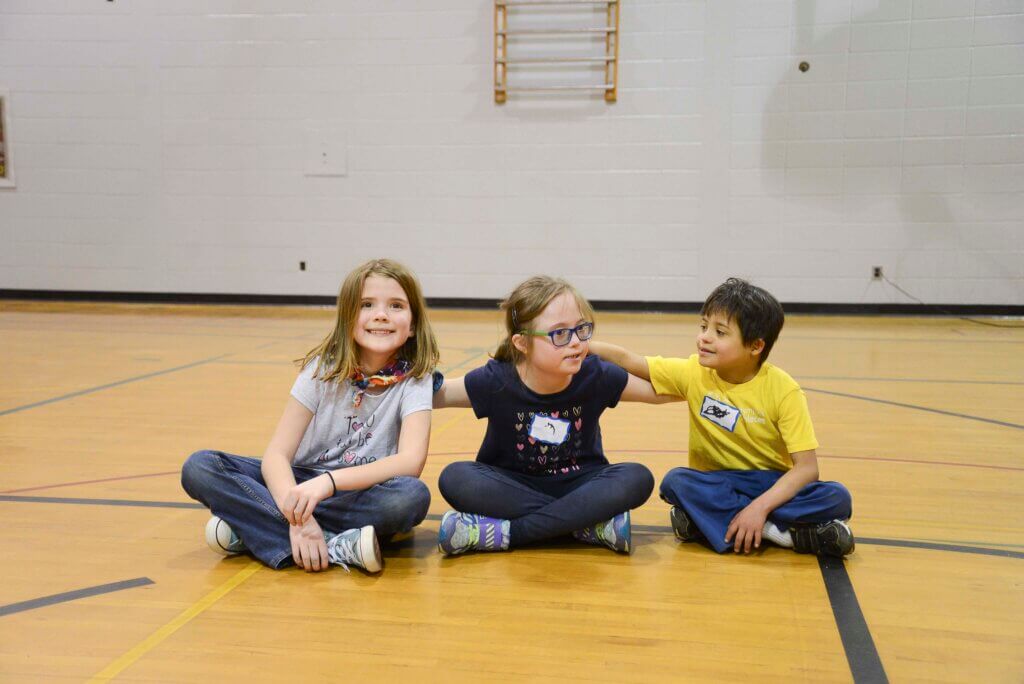Three smiling children sitting on a gym floor with their arms around each other.