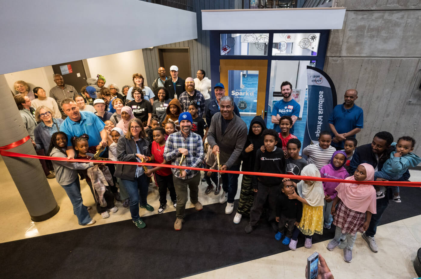 A group of people stand in front of the new Spark'd Studios location in Luxton Recreation Center. They are just about to cut a red ribbon.