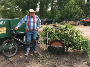 Man wearing a sunhat and garden knee pads standing in front of his bike with a trailer filled with yard waste.