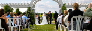 Picture of an outdoor wedding, the bride and groom are standing under an arch, while rows of guests are seated watching